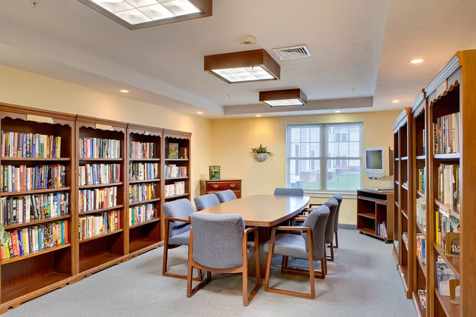 A well-lit room with wooden bookshelves filled with books lining both sides. In the center, there is a rectangular table surrounded by eight gray upholstered chairs. A window at the far end lets in natural light, and a small TV is placed on a stand near the window. The ceiling has recessed lighting and two large square light fixtures.