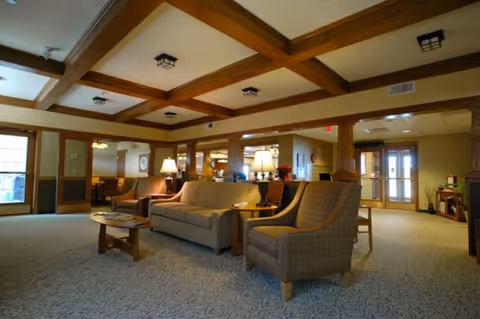 A spacious living room area in Windsor Manor featuring a beige sofa, two patterned armchairs, wooden coffee tables, and lamps on side tables. The ceiling has wooden beams, and the room is well-lit with natural light coming through windows and glass doors in the background.