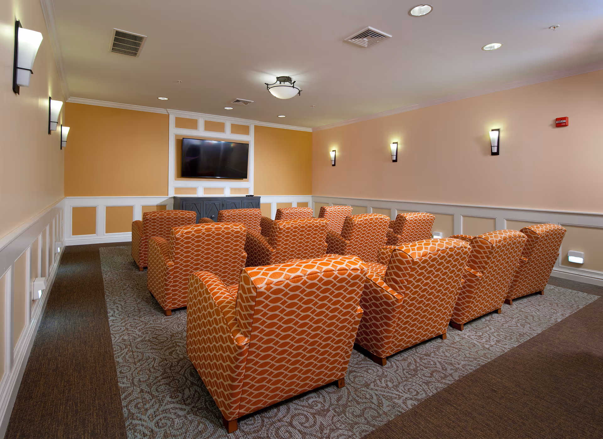 A small theater-style room with three rows of orange patterned armchairs facing a flat-screen TV mounted on a beige and white paneled wall. The room has soft lighting from wall sconces and ceiling lights, with carpeted flooring and a cozy atmosphere.