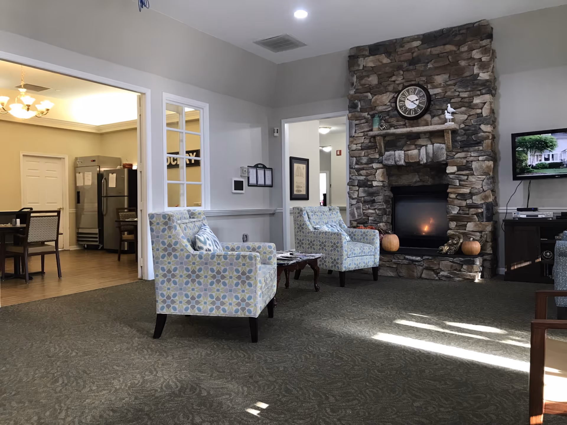 A cozy communal living room with patterned armchairs facing a stone fireplace and a TV, adjacent to a dining area.