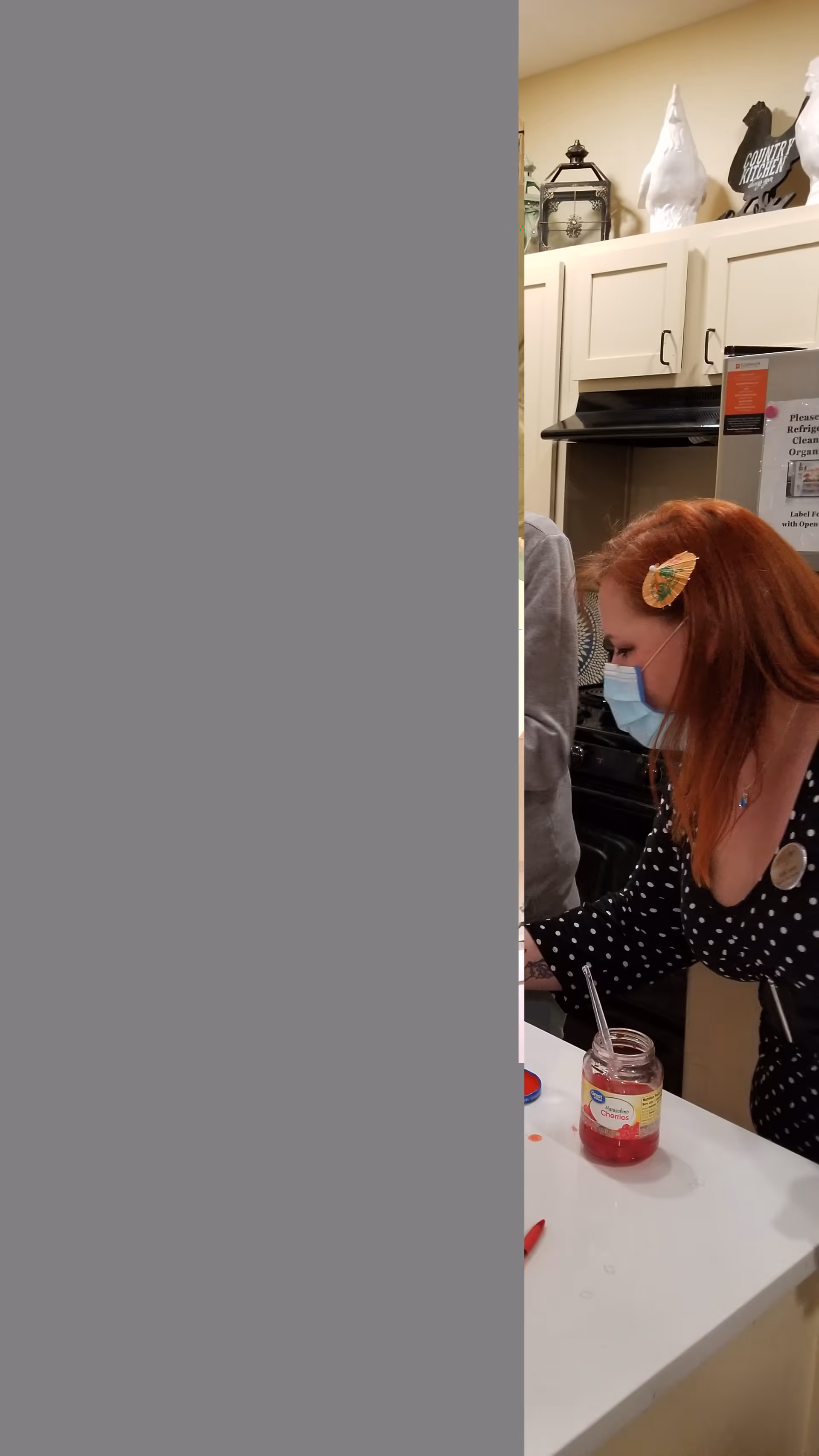 A woman wearing a face mask and a black polka dot dress with a butterfly hair clip is leaning over a kitchen counter. On the counter, there is an open jar of maraschino cherries with a spoon inside. The background shows white kitchen cabinets, a stove, and decorative items on top of the cabinets.