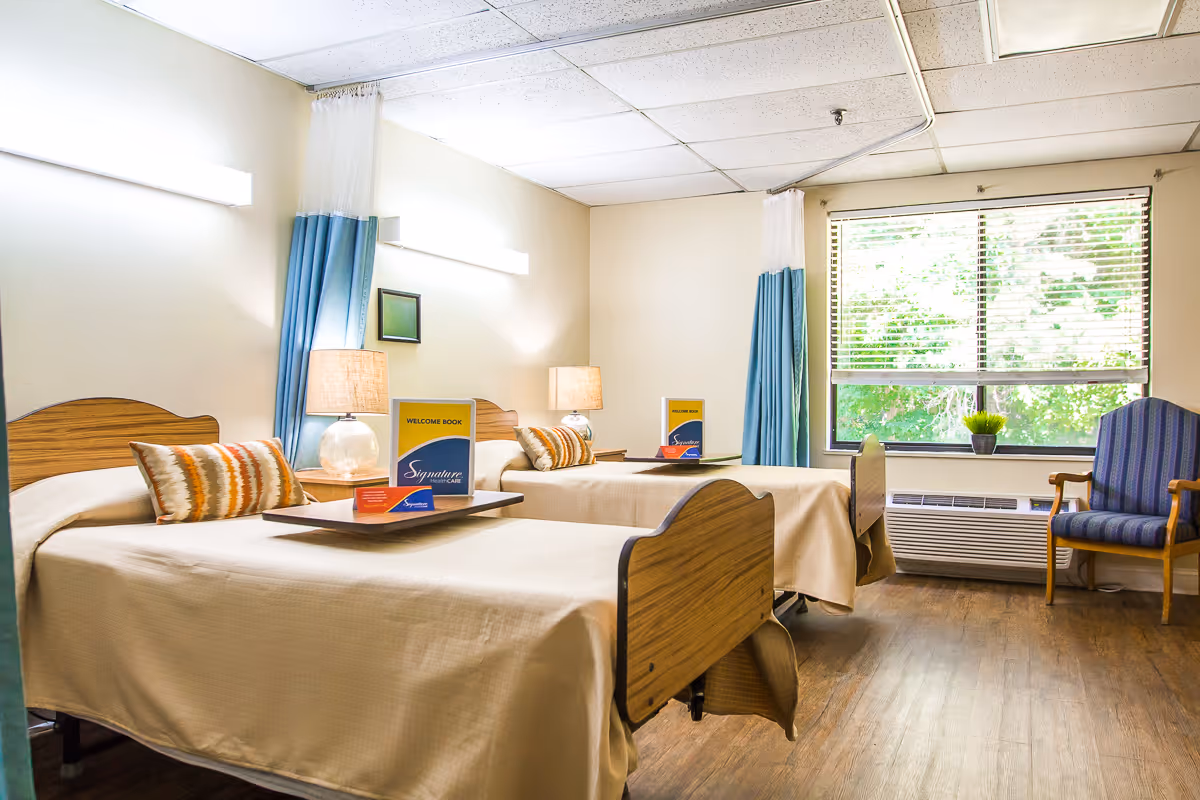 A bright senior care room with two single beds, bedside tables and lamps, privacy curtains, and a window overlooking greenery.