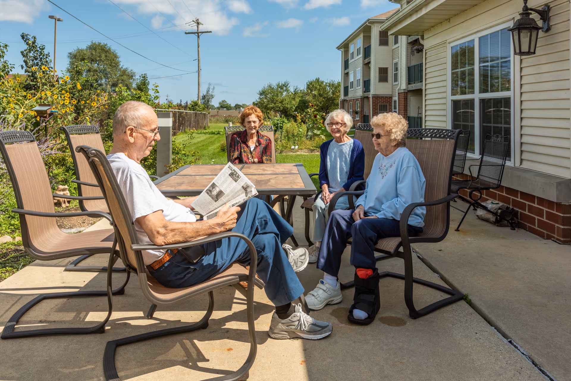 Four elderly people sitting outside on patio chairs around a table. One man is reading a newspaper while three women are sitting and chatting. The setting is a sunny day with a garden and a building in the background.