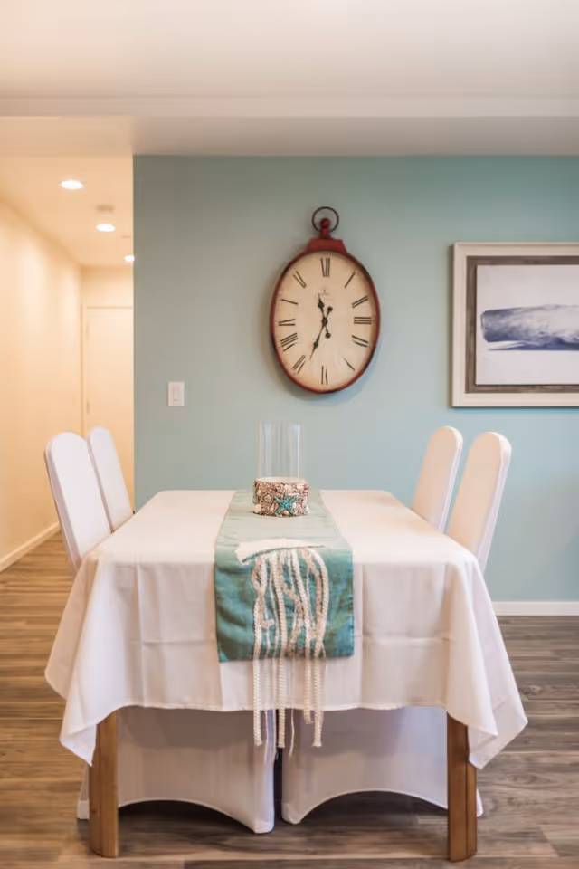 A dining table covered with a white tablecloth and a teal table runner with decorative tassels, surrounded by four white chairs. On the wall behind the table is a large vintage-style clock and a framed artwork. The room has light blue walls and wood flooring.