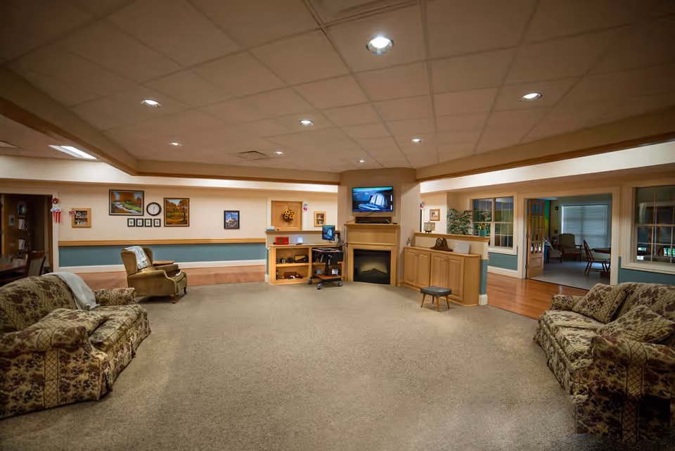 A spacious living room area in a senior living facility featuring two floral patterned sofas, a green armchair, a small stool, and a fireplace with a mounted TV above it. The walls are decorated with framed pictures and a clock. There is a computer workstation next to the fireplace and large windows and doorways leading to adjacent rooms.