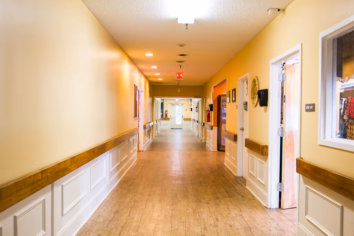 A long, well-lit hallway in a senior living facility with light yellow walls, wooden handrails, and light wood flooring. Several doors and windows line the hallway, and there is an exit sign at the far end above a door.