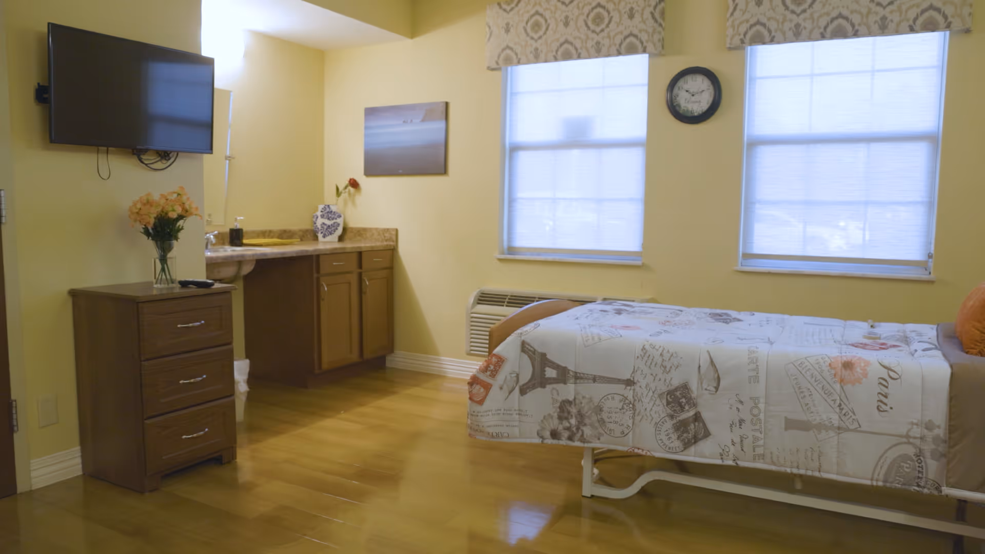 A bright and clean bedroom in a senior living facility with a single bed covered in a Paris-themed bedspread, two large windows with blinds and patterned valances, a wall-mounted TV, a wooden dresser with a vase of flowers, and a small sink with cabinets underneath.