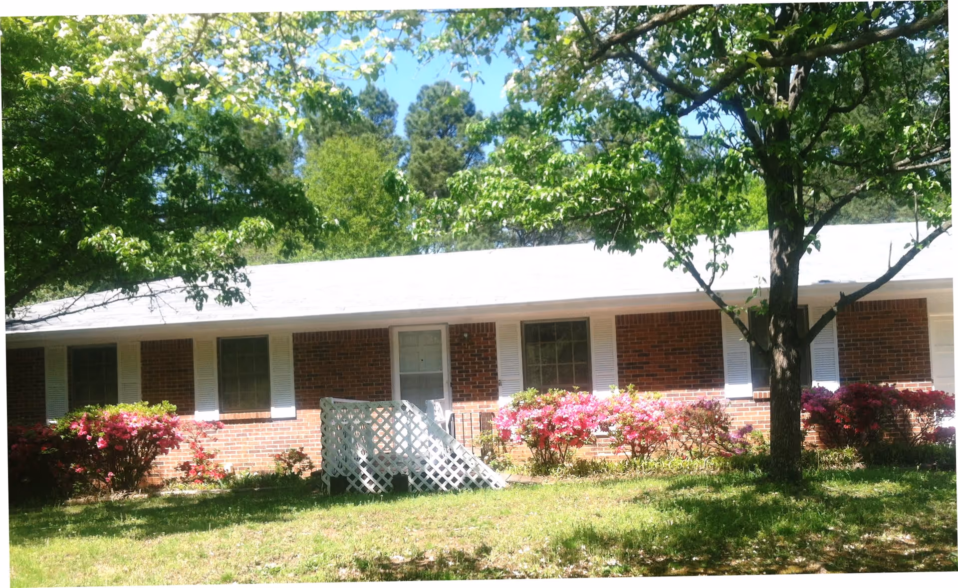 Single-story brick building with white shutters and a white roof, surrounded by green trees and bushes with pink flowers, under a clear blue sky.