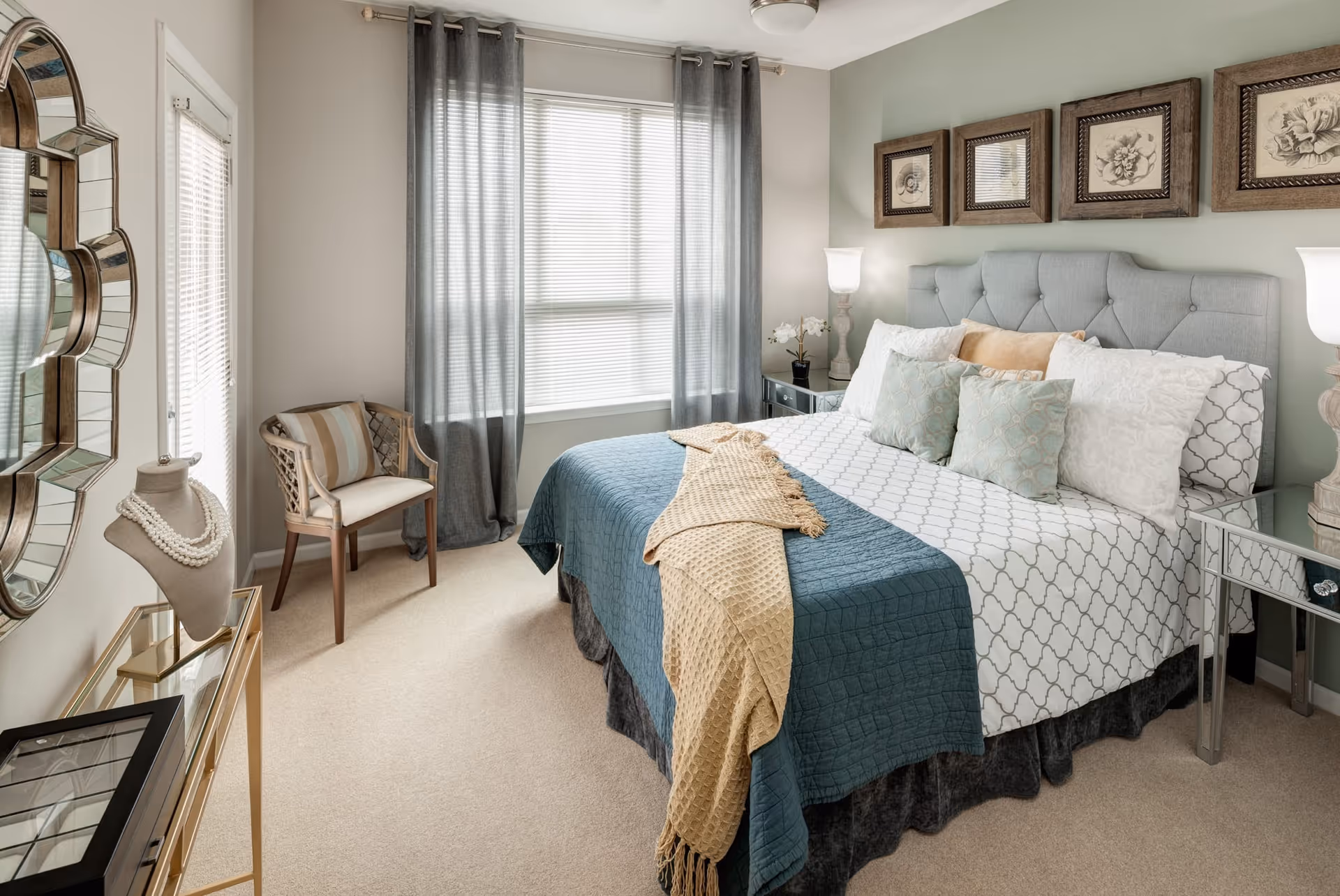 A neatly arranged bedroom with a large bed featuring a tufted headboard, multiple pillows, and a blue and beige throw blanket. There are two mirrored nightstands with lamps on either side of the bed. A window with gray curtains allows natural light into the room. A decorative chair with a striped cushion sits near the window, and a unique wall mirror and jewelry display are visible on the left side.