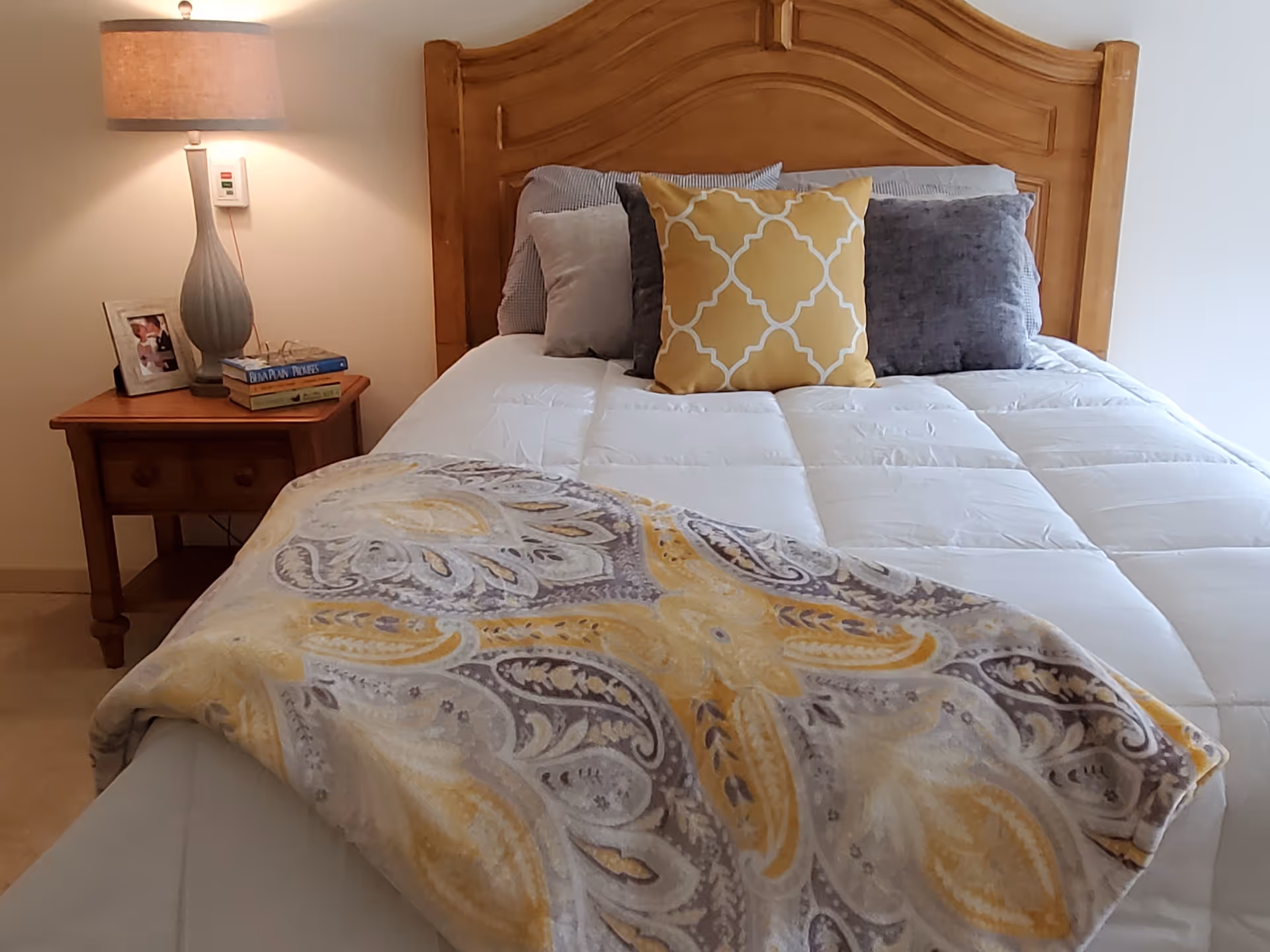 A neatly made bed with a wooden headboard, white comforter, and multiple pillows including a yellow decorative pillow. A patterned blanket with yellow and gray designs is draped over the foot of the bed. To the left of the bed is a wooden nightstand with a lamp, a framed photo, and a few books.