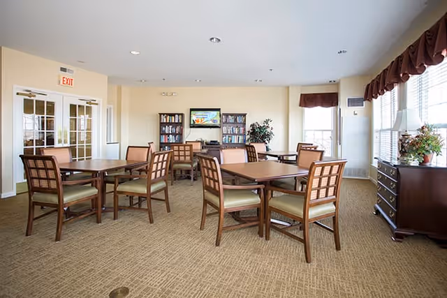 A well-lit common area with multiple wooden tables and chairs arranged on a carpeted floor. There are large windows with brown valances allowing natural light to fill the room. A wooden dresser with a lamp and plant sits near the windows. In the background, there are bookshelves with books and a television mounted on the wall. The walls are painted a light cream color.