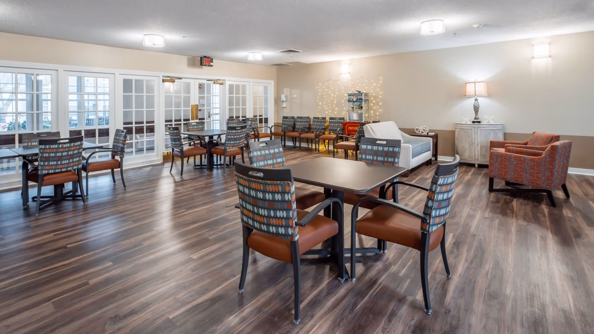 A spacious common area with several tables and chairs arranged on a wood floor. The room features a white sofa, an orange armchair, a popcorn machine against the wall, and soft lighting from wall-mounted fixtures and a table lamp. Large glass doors lead to another room with more seating visible.