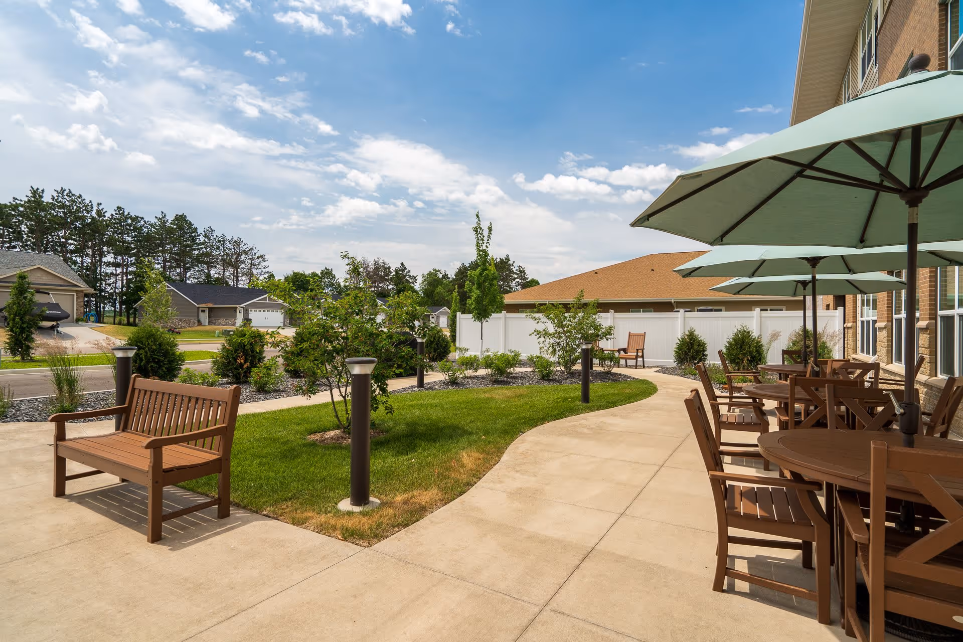 Outdoor patio area at Caretta Senior Living - Holmen featuring wooden benches, tables with chairs, and large green umbrellas. The patio is surrounded by a well-maintained lawn, small trees, and shrubs, with a clear blue sky overhead and residential houses visible in the background.