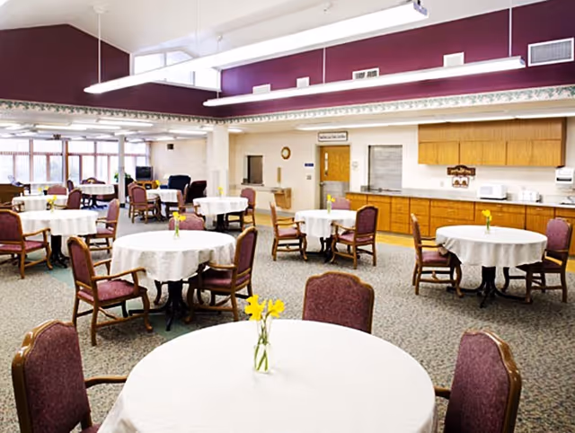 A spacious dining room with round tables covered in white tablecloths, each table having a small vase with yellow flowers. The room has maroon walls near the ceiling, large windows letting in natural light, and wooden cabinets along one wall. Several chairs with maroon upholstery are arranged around the tables.