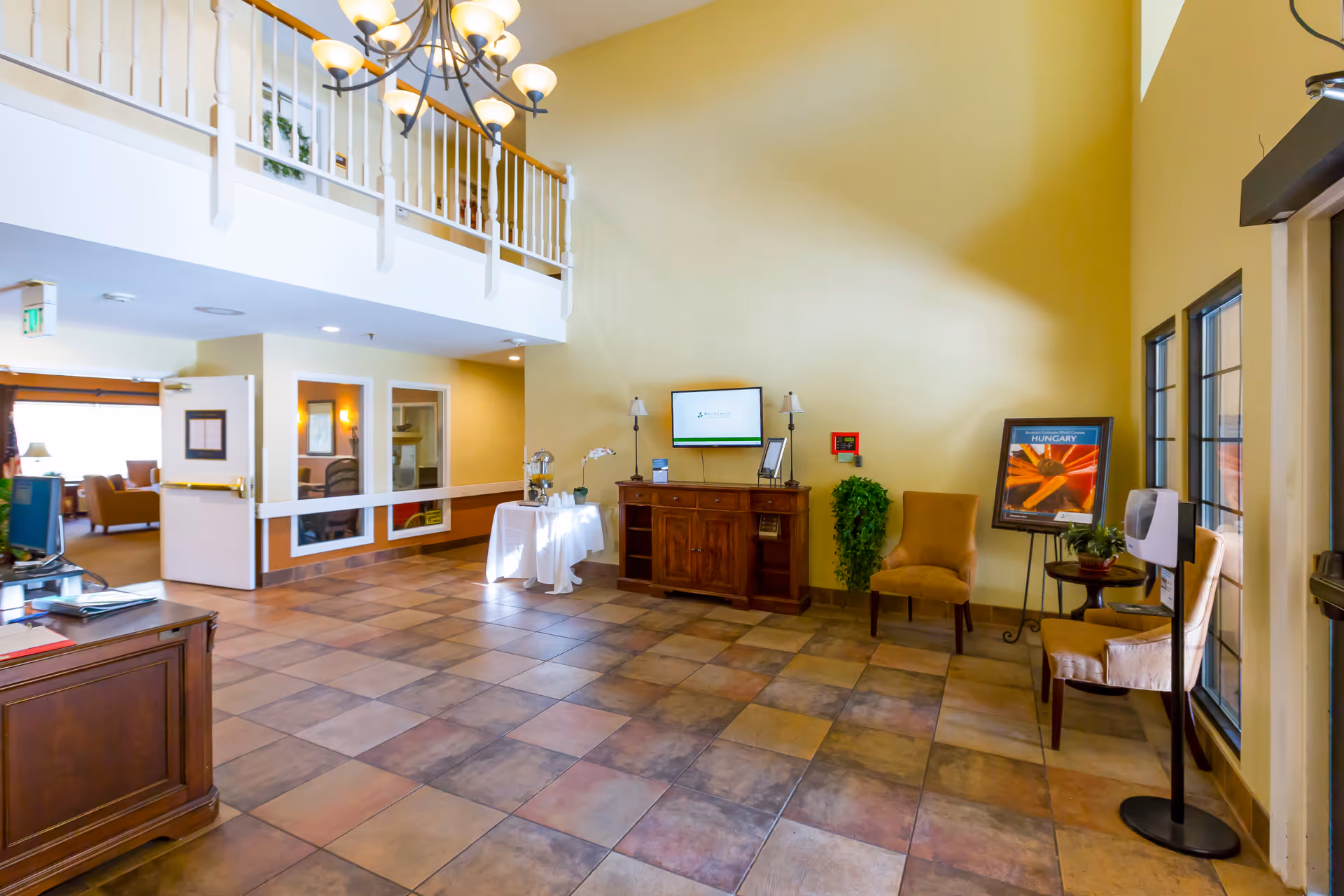 A spacious, well-lit interior area with a high ceiling and tiled floor. There is a wooden cabinet against a yellow wall with a TV mounted above it, flanked by two lamps. Two upholstered chairs and a small table with a plant are positioned near windows on the right. A chandelier hangs from the ceiling, and an open doorway leads to another room with seating visible. A small table with a white tablecloth and water dispenser is also present.