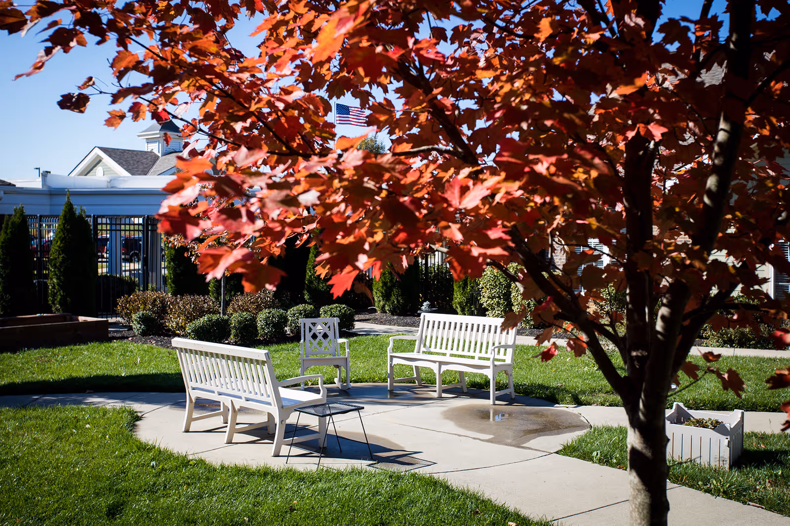 Outdoor seating area with white benches and chairs arranged on a circular concrete patio surrounded by green grass and landscaping. A tree with red autumn leaves partially shades the area. In the background, there is a fence, shrubs, and a building with an American flag flying.
