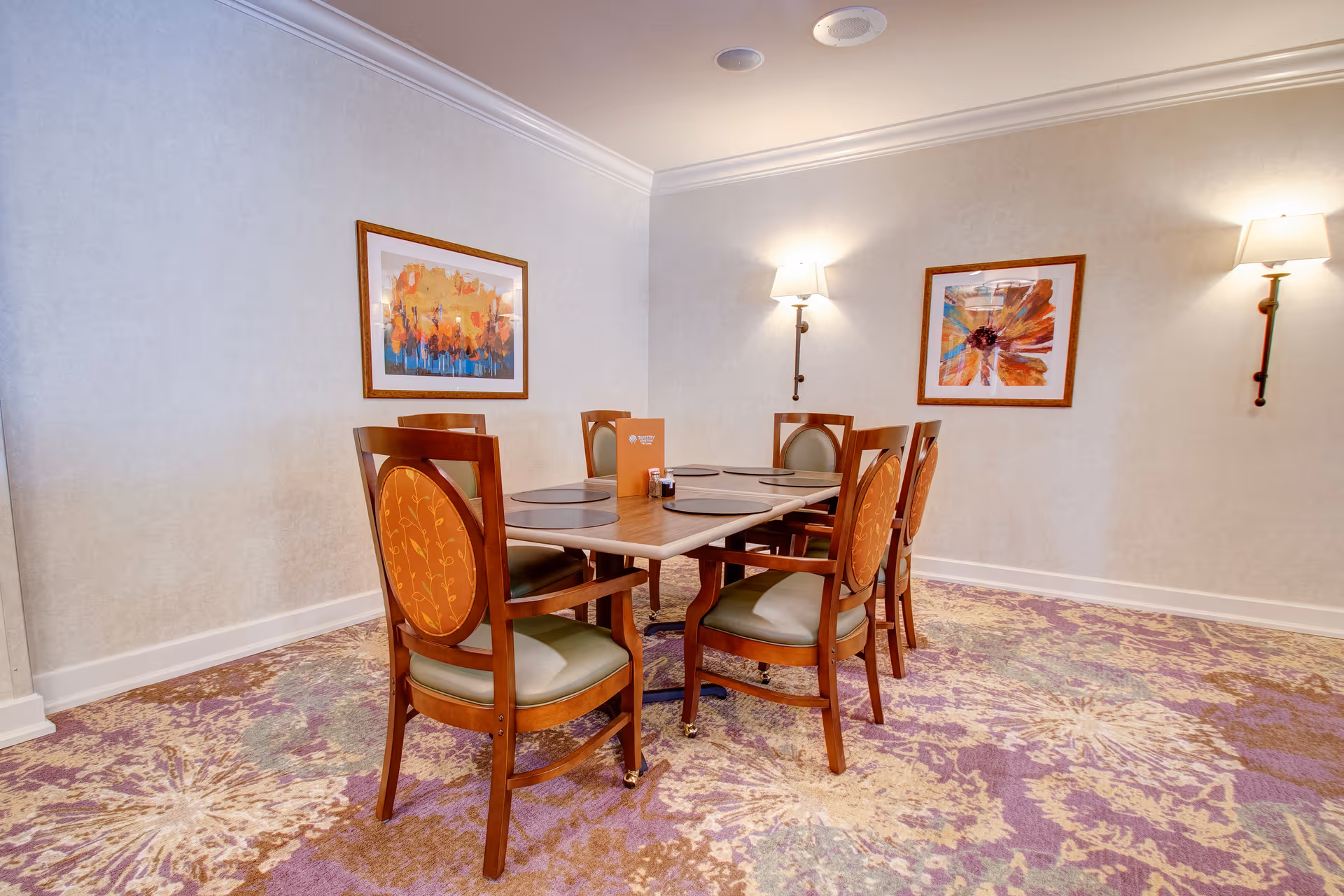A dining area with a rectangular wooden table surrounded by six wooden chairs with cushioned seats and backs. The floor is covered with a patterned carpet in shades of purple, beige, and green. Two framed abstract paintings hang on the light-colored walls, and two wall-mounted lamps provide soft lighting.