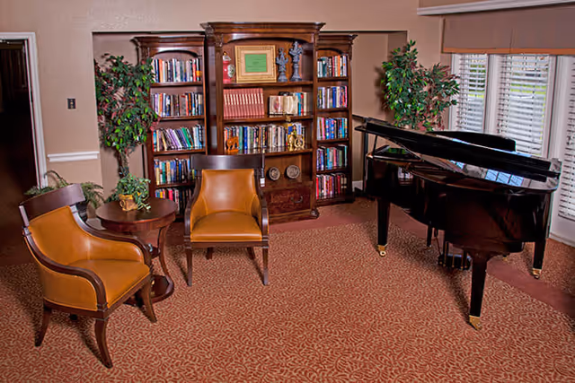 A cozy living room area featuring two brown leather chairs with wooden frames around a small round wooden table with a plant on it. Behind the chairs is a large wooden bookshelf filled with books and decorative items. To the right is a black grand piano near windows with white blinds and beige valances. Two tall green plants are placed on either side of the bookshelf.