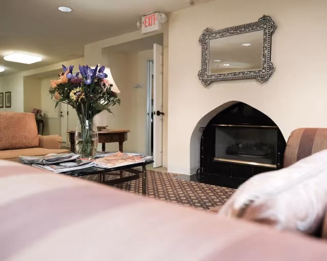 Cozy seating area in a senior living lounge with couches, a coffee table holding magazines and a vase of flowers, and a fireplace beneath a decorative mirror.