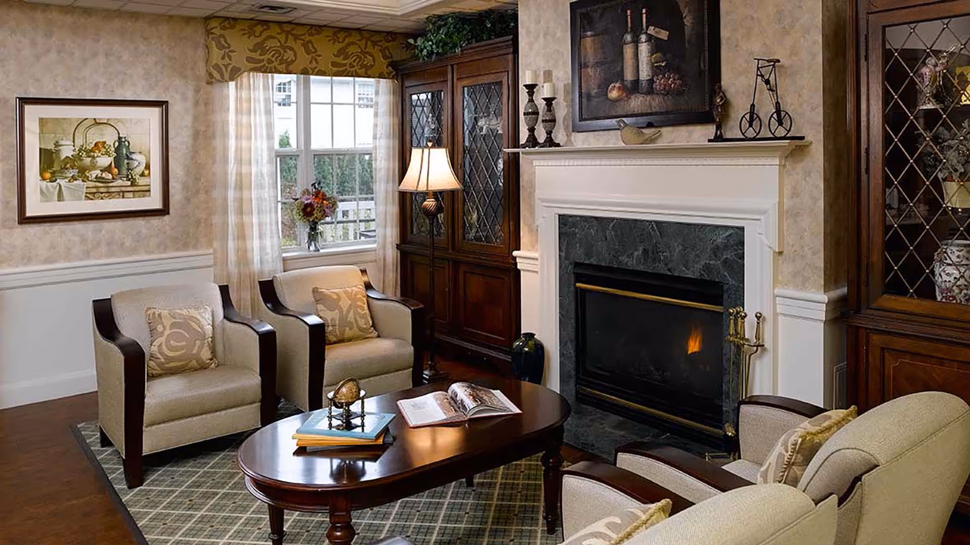 A cozy living room with a lit fireplace surrounded by a white mantel and dark marble. The room features four beige armchairs with dark wooden arms, a wooden coffee table with books and a decorative globe, a floor lamp, a window with sheer curtains, and two wooden display cabinets with glass doors. The walls are decorated with framed artwork and patterned wallpaper.