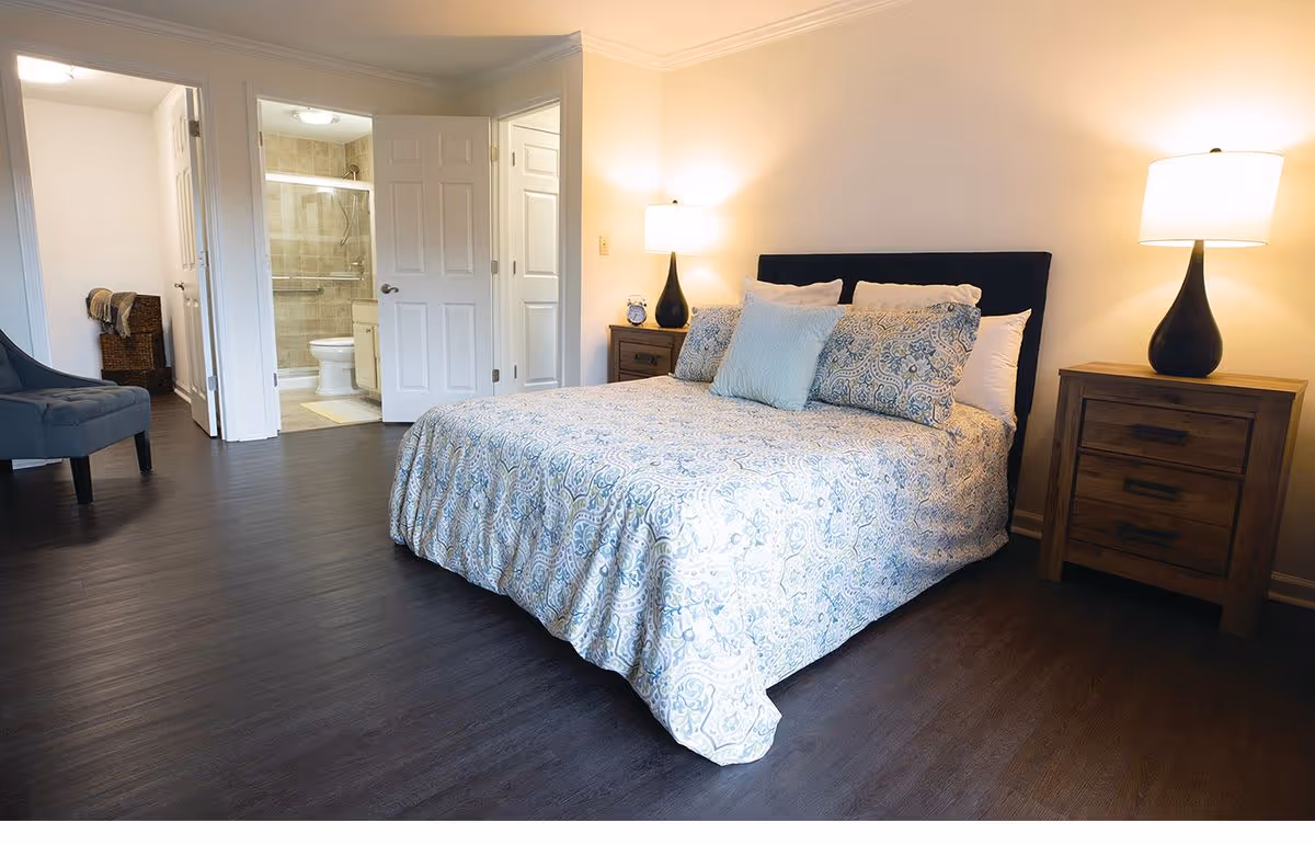 Well-lit bedroom with a patterned bedspread, two nightstands and lamps, and an open door revealing an en-suite bathroom.