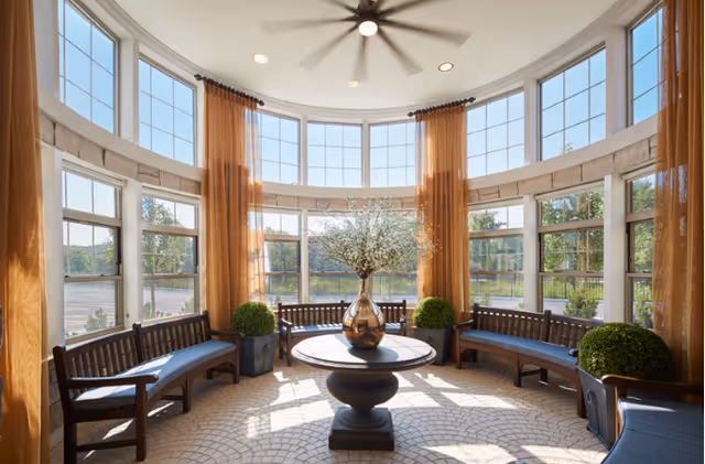 Circular glass-walled sunroom with curved benches, orange drapes, and a central vase on a pedestal.