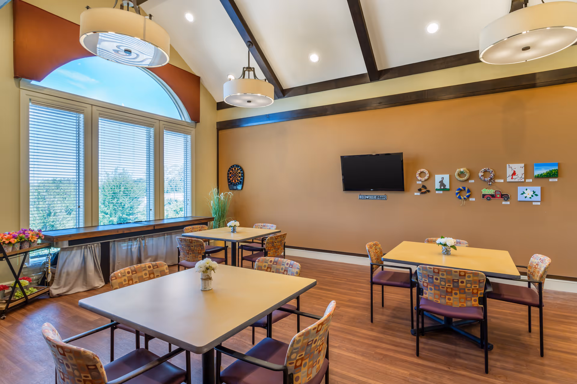 A bright and spacious common room with high vaulted ceilings featuring dark wooden beams. The room has large windows with blinds and an arched top, allowing natural light to fill the space. There are three square tables, each surrounded by four patterned chairs with purple seats. Small flower arrangements are placed on each table. On the far wall, a flat-screen TV is mounted above a sign, and various small artworks and wreaths are displayed. A dartboard is mounted on the left wall near a tall plant. The floor is wooden, and two large round ceiling lights hang from the ceiling.