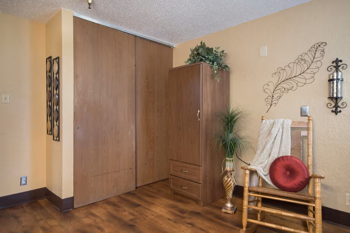 A cozy corner of a room featuring a wooden rocking chair with a red round cushion and a white throw blanket draped over it. Next to the chair is a tall wooden cabinet with two drawers and a green plant on top. The wall behind has decorative metal wall art and a candle holder. The floor is wooden, and there is a closet with sliding wooden doors.
