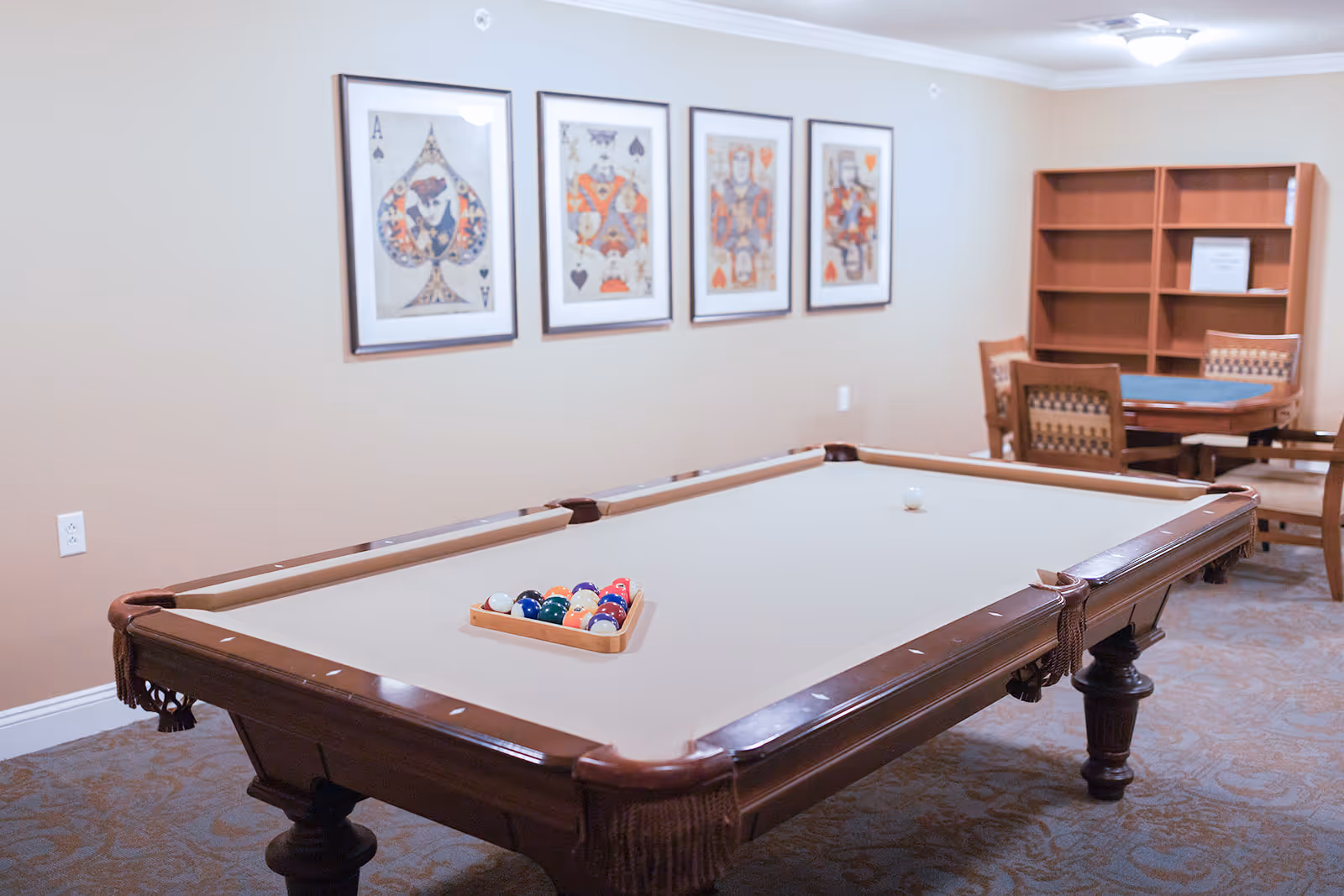 A game room with a beige pool table set up for a game, featuring a rack of colorful billiard balls and a white cue ball. On the wall behind the pool table are four framed playing card artworks. In the background, there is a wooden table with chairs and an empty bookshelf. The room has beige walls and carpeted flooring.