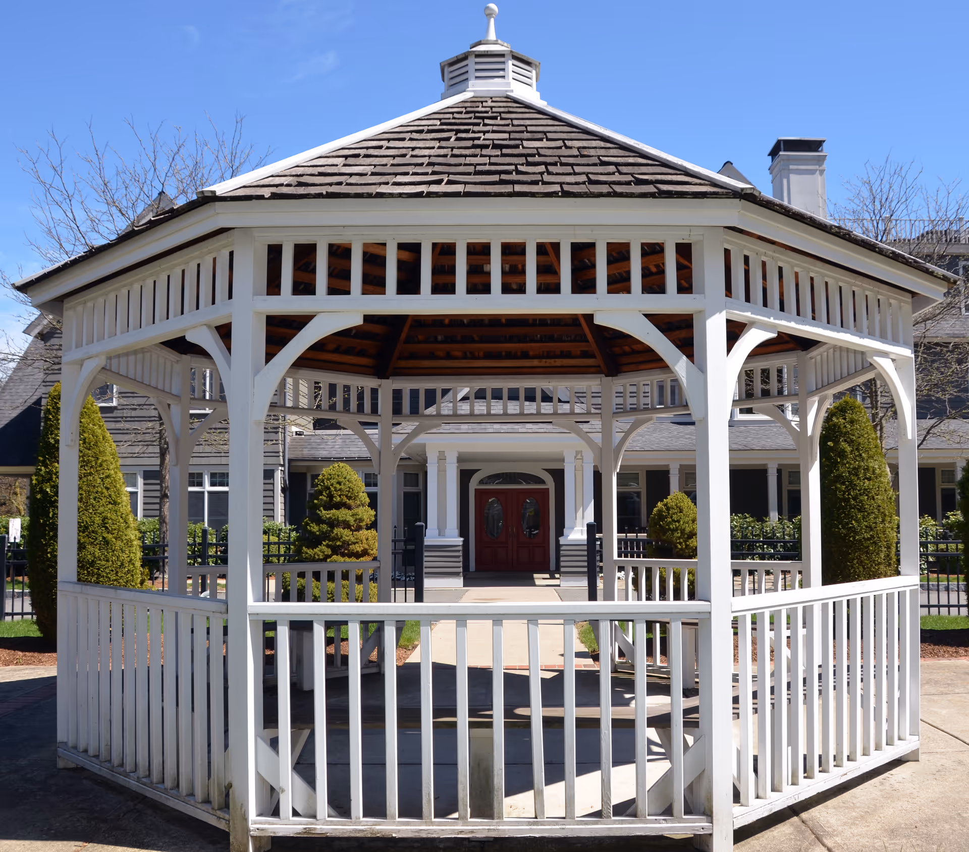 A white wooden gazebo sits in front of a building entrance with red double doors, trimmed shrubs, and a clear blue sky.