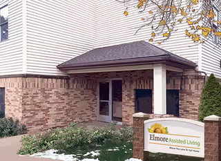Exterior view of Elmore Assisted Living & Memory Care building entrance with brick and siding walls, a small covered porch, and a sign in front surrounded by greenery and some fallen leaves.