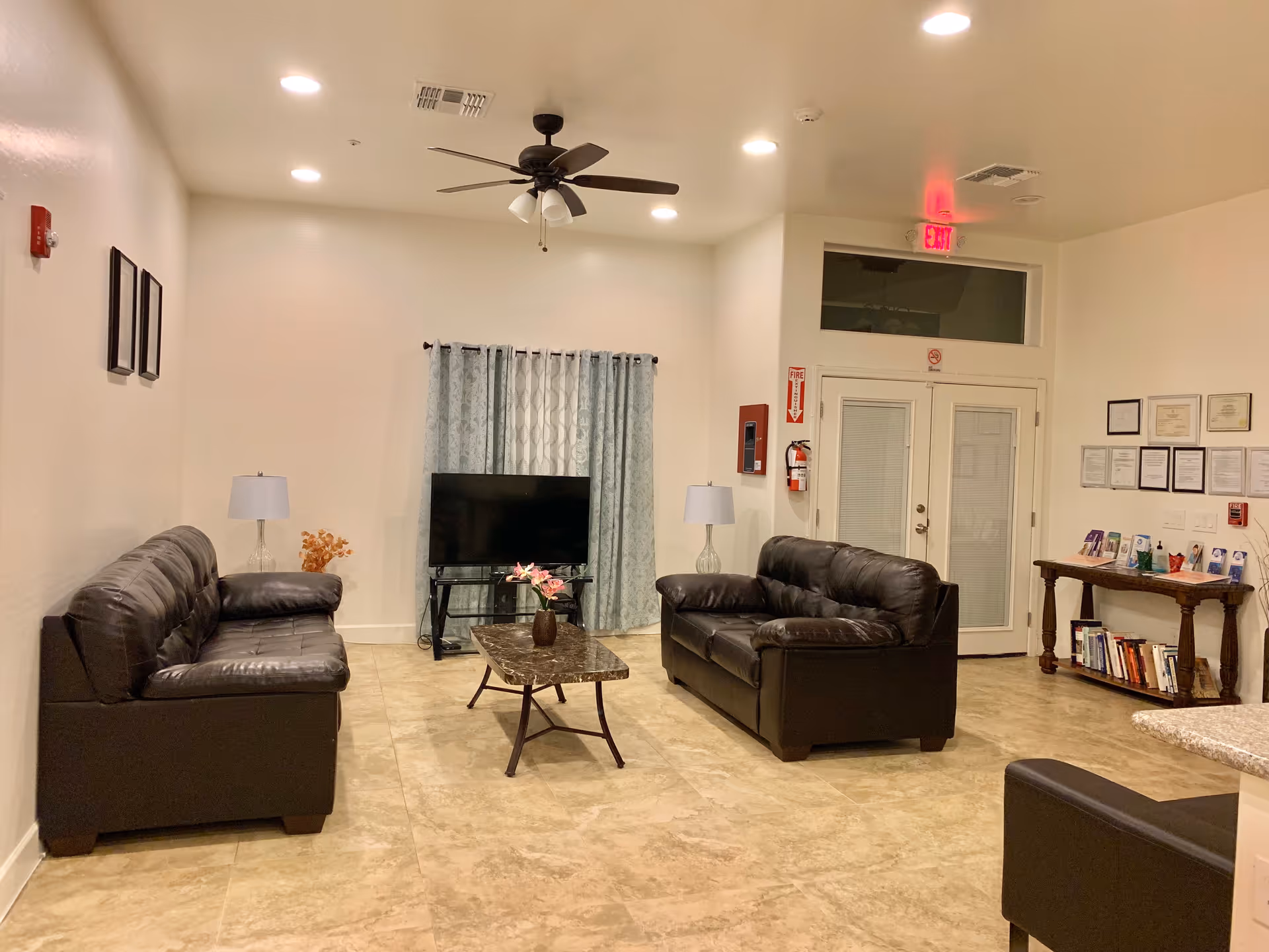 A cozy living room area with two black leather sofas facing each other, a marble coffee table with a vase of flowers in the center, a flat-screen TV on a stand against the wall with blue patterned curtains behind it, two table lamps on either side of the sofas, a ceiling fan with lights, and a small wooden table with books and pamphlets near a set of double doors.