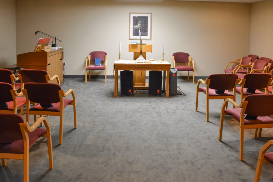 A small chapel or meditation room with rows of wooden chairs with red cushions arranged facing a wooden altar table. The altar has two candlesticks, a microphone, and a framed picture on the wall behind it. There is a wooden organ or piano on the left side of the room.