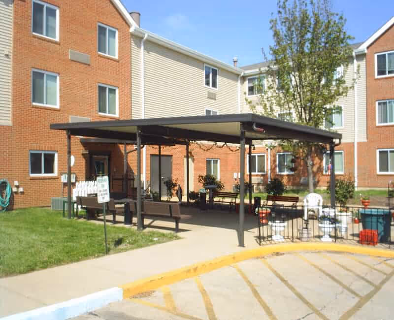 Outdoor covered seating area with benches and chairs in front of a three-story brick and siding apartment building under a clear blue sky.