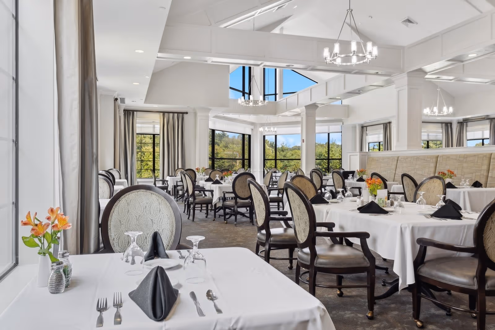 Bright and elegant dining room with multiple tables covered in white tablecloths, each set with black folded napkins, glassware, and silverware. The room features large windows with views of greenery outside, chandeliers hanging from a high ceiling with exposed beams, and upholstered chairs around the tables. Fresh flowers in vases decorate the tables.