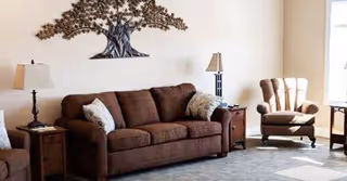 A cozy living room with a brown sofa adorned with two light-colored pillows, a cushioned armchair, two side tables each with a lamp, and a decorative metal tree wall art above the sofa. Sunlight streams in from a window on the right side.