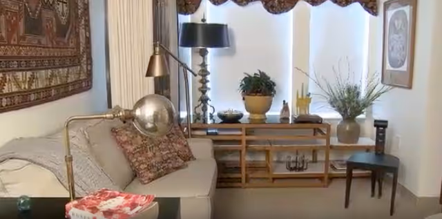 A cozy living room area with a beige sofa adorned with patterned pillows and a throw blanket. A brass floor lamp with a spherical shade is positioned next to the sofa. Behind the sofa, there is a wooden console table with various decorative items including potted plants, a black table lamp, and small sculptures. The room has light-colored walls with framed artwork and a decorative tapestry hanging on one side. A small dark wooden chair is placed near the window with sheer white curtains.