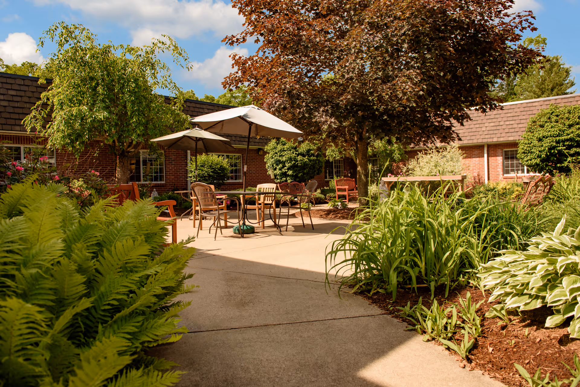 Outdoor courtyard area with patio tables and chairs under umbrellas surrounded by lush green plants, trees, and shrubs. The background shows a brick building under a partly cloudy blue sky.