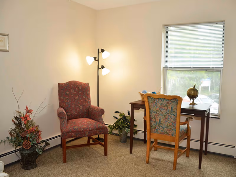 A cozy corner of a room with two upholstered chairs, one red patterned armchair and one floral patterned wooden chair, positioned near a small wooden desk with a globe on it. A floor lamp with three lights stands behind the red chair, and there are two potted plants on the floor near the walls. A window with blinds is visible behind the desk.