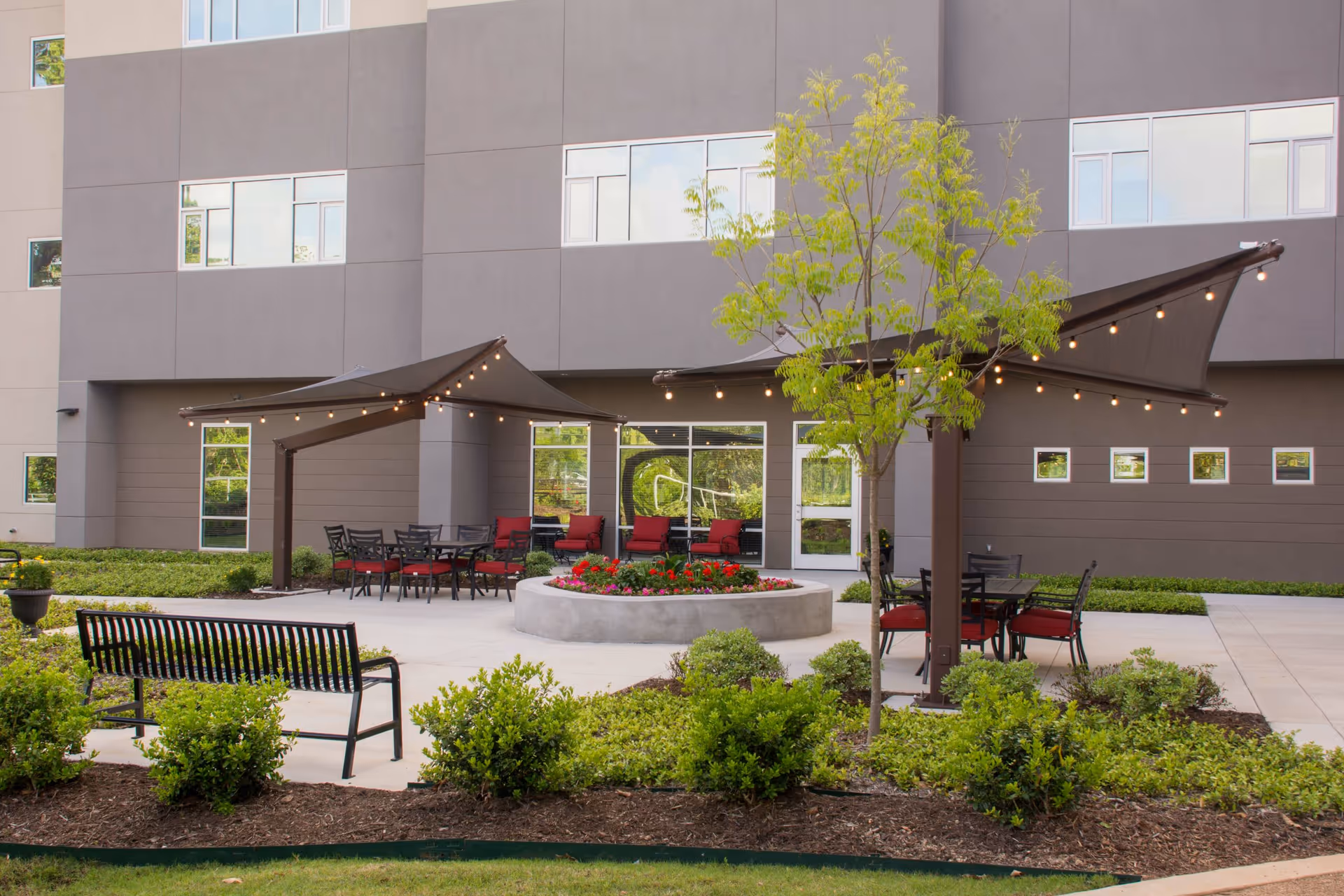 Outdoor patio area at a senior living facility with a circular flower bed in the center, surrounded by seating including black metal benches and tables with red cushioned chairs. Two large triangular shade sails with string lights are installed above the seating areas. The building's exterior wall with multiple windows is visible in the background.