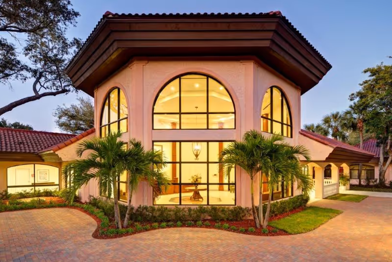 Exterior view of a building at dusk with large arched windows illuminated from inside, surrounded by palm trees and landscaped greenery, with a paved driveway in front.