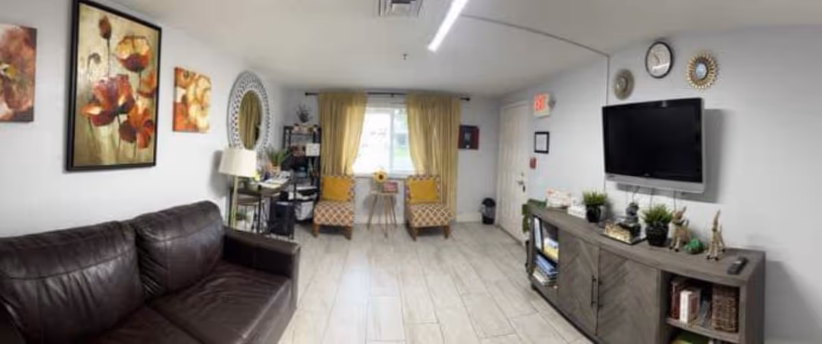 Cozy common living room with a dark leather sofa, a wall-mounted TV above a console, two patterned armchairs by a curtained window, and decorative wall art.