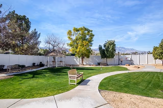 Outdoor garden area with a curved concrete pathway, green grass, several benches, a white gazebo, and trees under a blue sky with mountains in the background, enclosed by a white fence.