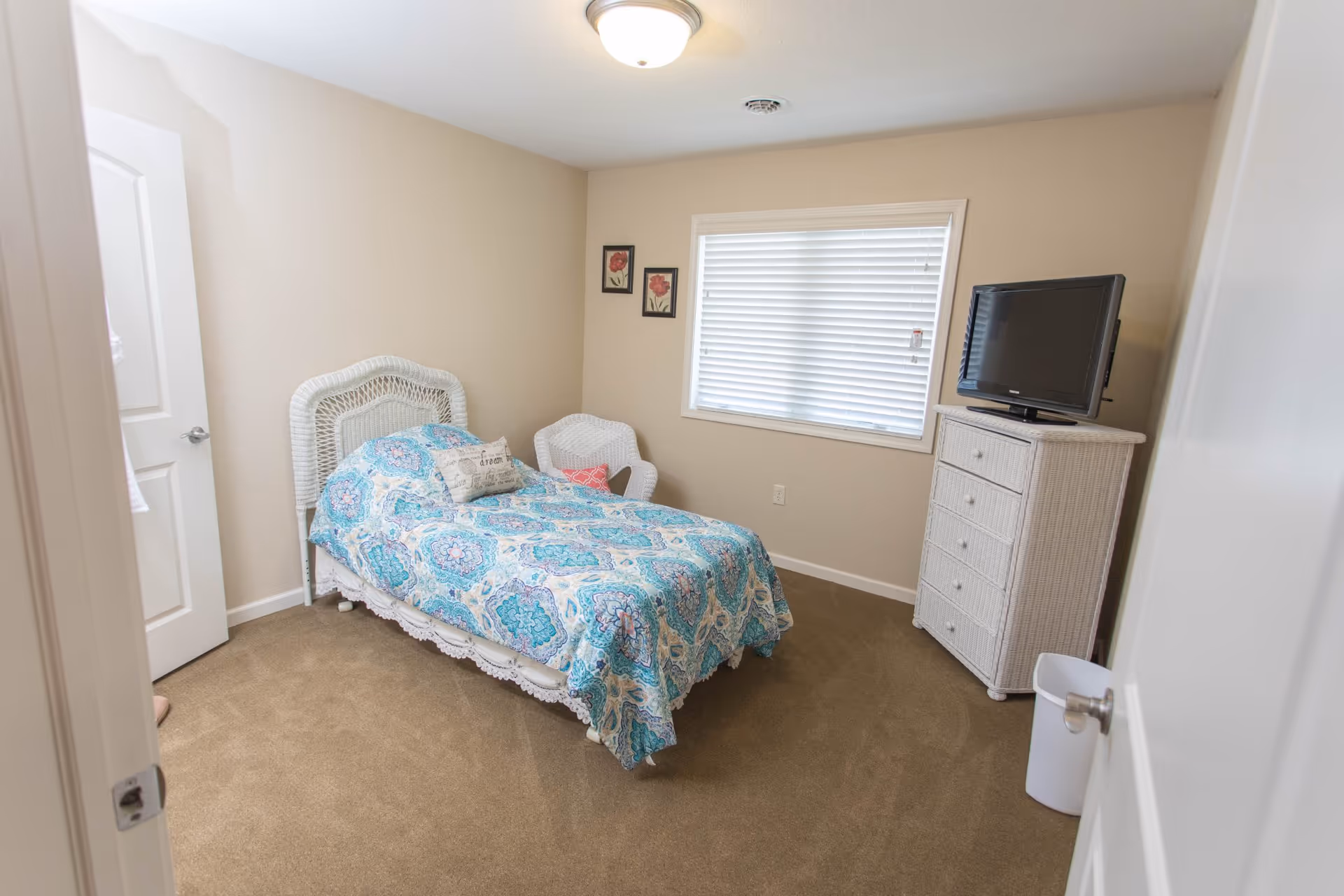 A small bedroom with beige walls and carpet. The room features a single bed with a blue and white patterned bedspread, a white wicker headboard, and a decorative pillow. There is a white wicker chair in the corner and a white wicker dresser with a flat-screen TV on top. A window with closed blinds is on the wall behind the dresser, and two framed floral pictures hang on the adjacent wall.