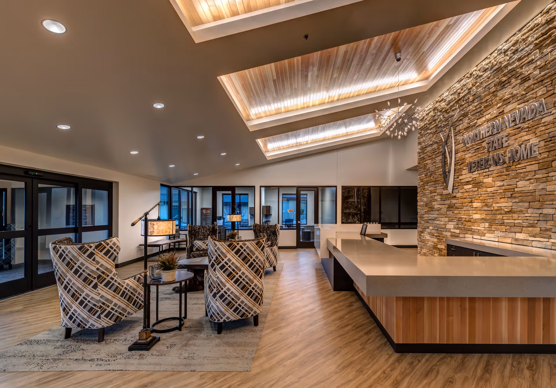 Interior view of a modern reception area at Northern Nevada State Veterans Home featuring a stone accent wall with the facility's name, a large reception desk, patterned armchairs arranged around small tables, wooden flooring, and recessed ceiling lighting.