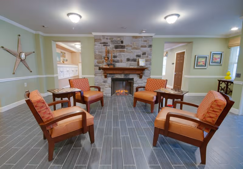 A cozy seating area in a senior living facility with four cushioned armchairs arranged around two small tables, each with a chessboard. The room features a stone fireplace with a wooden mantel, light green walls with white trim, and framed artwork. The floor is covered with gray tiles, and there are ceiling lights providing illumination.