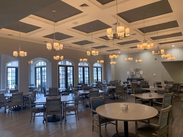 A spacious dining room with multiple tables and chairs arranged neatly. The room features large arched windows allowing natural light to enter, and several modern chandeliers hanging from a coffered ceiling. The floor is wooden, and the overall ambiance is warm and inviting.