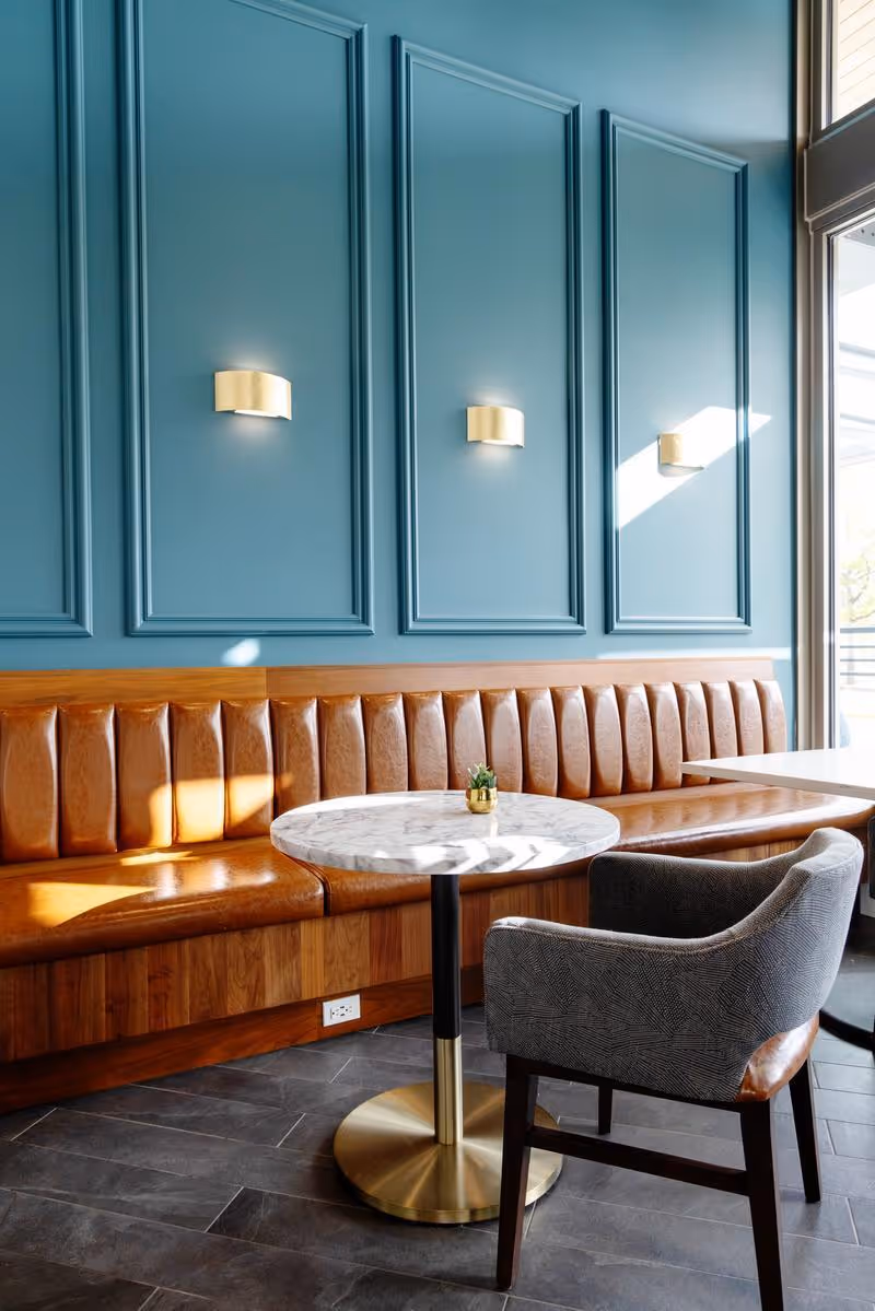 A cozy seating area with a long brown leather bench against a teal paneled wall featuring three gold wall sconces. In front of the bench is a round marble table with a small potted plant, accompanied by a single upholstered chair. Natural light streams in from a large window on the right.
