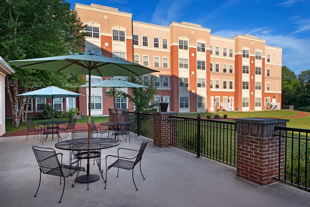 Outdoor patio area with metal tables and chairs under large umbrellas, adjacent to a multi-story residential building with many windows. The patio is surrounded by a black metal fence with brick pillars, and there is a grassy lawn and trees in the background under a clear blue sky.
