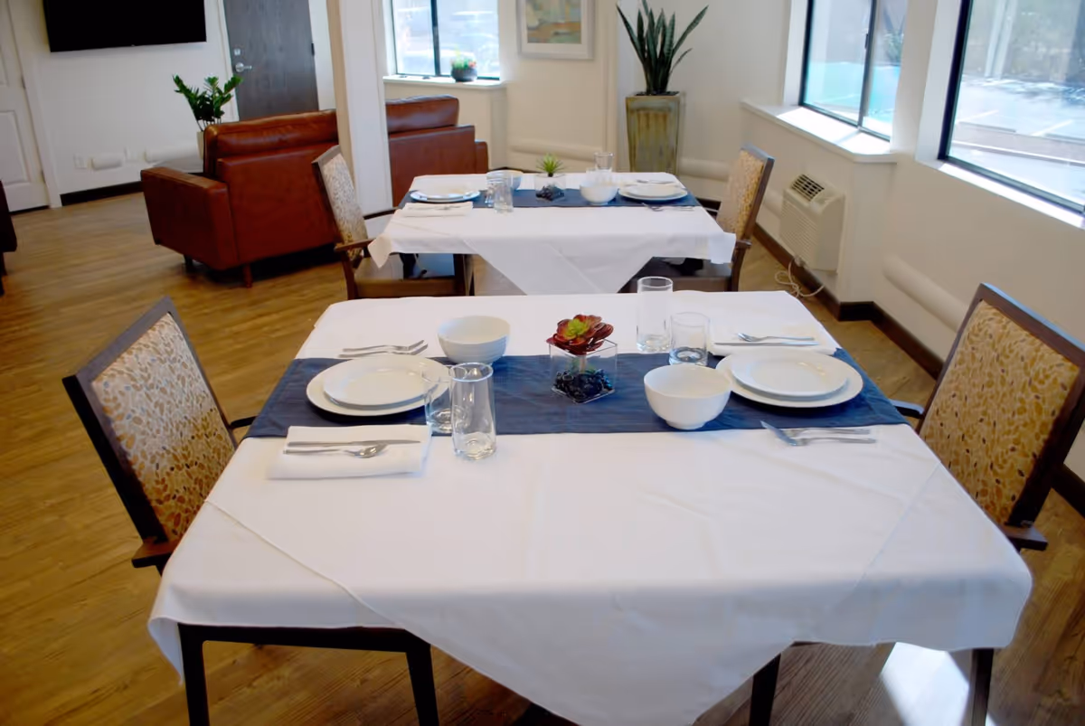 Two dining tables set with white tablecloths, blue runners, plates, bowls, glasses, and silverware in a bright room with large windows. There are upholstered chairs around the tables, a potted plant in the corner, and a seating area with brown leather sofas in the background.